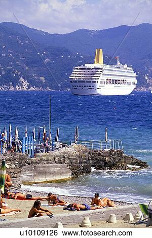Stock Photograph - Sunbathers and umbrellas rocky waters edge with cruise ship in bay. Fotosearch