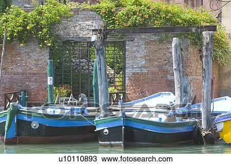 Stock Image - Blue barges in sunlight moored at canal side with red brick wall covered with vines. Fotosearch