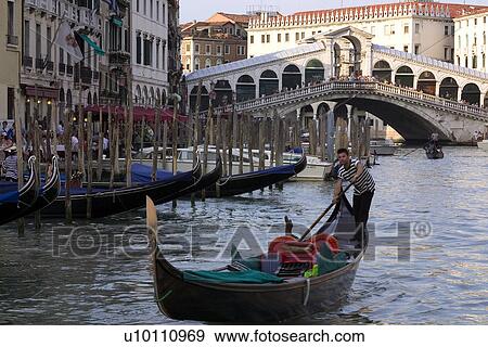 Stock Photo - Gondolier in Gondola, other gondolas moored at canal side posts and Rialto Bridge in afternoon sun. Fotosearch