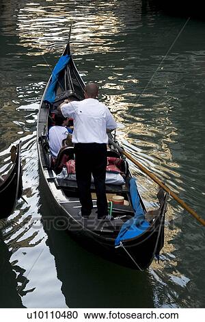 Stock Image - Gondolier in Gondola with passengers on quiet canal - reflections in water. Fotosearch