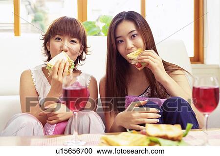 Stock Image - Two Young Adult Women Eating Sandwiches and Drinking Red Wine, Looking at Camera, Front View. Fotosearch
