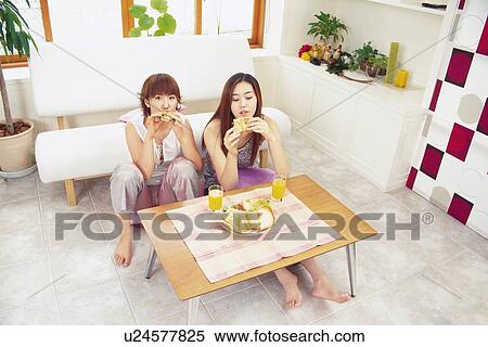 Stock Photography - Two Young Adult Women Eating Sandwiches in a Living room, High Angle View, Looking at Camera. Fotosearch