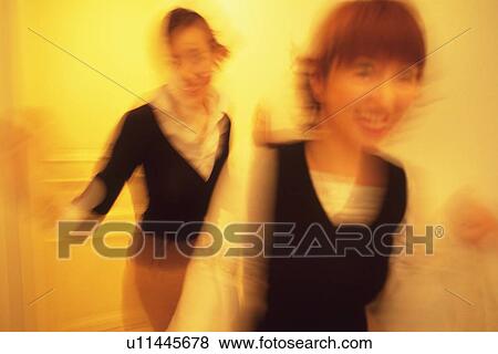Stock Photo - Two Young Adult Women Walking Together in a Hallway, Smiling, Blurred Motion, Front View. Fotosearch