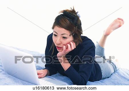 Stock Image - Young Adult Woman Operating her Laptop, Lying on her Bed Side View, High Angle View. Fotosearch
