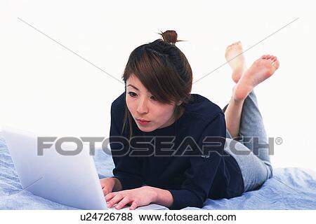 Stock Photo - Young Adult Woman Operating her Laptop, Lying on her Bed, High Angle View, Side View. Fotosearch