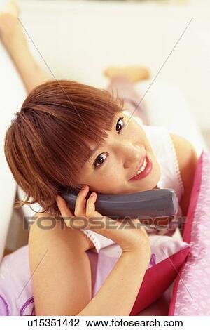 Stock Image - Young Adult Woman Talking on the Phone Smiling While Laying on a Couch, Looking at Camera, High Angle View Differential Focus. Fotosearch