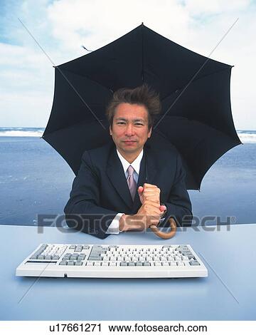 Stock Image - Image of a Businessman Sitting In Front of a Keyboard While Putting Up an Umbrella at the Beach, Looking at Camera, Front View. Fotosearch