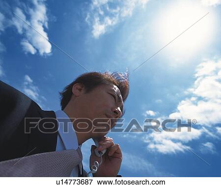 Stock Photo - Image of a Businessman Talking on a Phone Under a Blue Sky and the Sun Shining Brightly, Low Angle View. Fotosearch