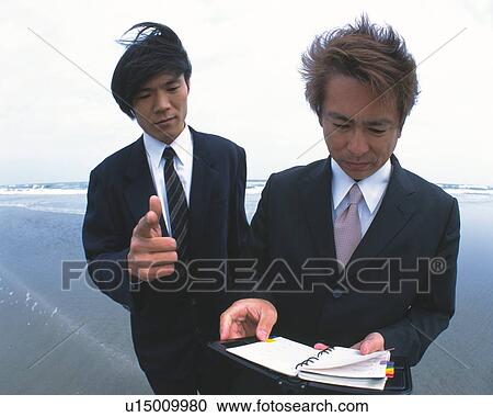 Stock Image - Image of Two Businessmen Looking at an Appointment Book at the Beach, Looking Down, Front View. Fotosearch
