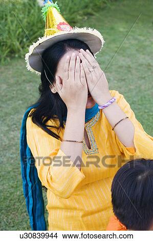 Picture - Close-up of a mid adult woman wearing a birthday hat and covering her face with her hands. Fotosearch
