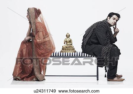 Stock Photo - Side profile of a newlywed couple sitting on opposite ends of a bench with a statue of Buddha between them. Fotosearch