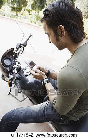 Stock Image - Close-up of a young man sitting on a motorcycle and using a mobile phone. Fotosearch