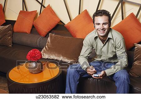 Portrait of a young man sitting on a couch and holding a mobile phone View Large Photo Image Stock Image - Portrait of a young man sitting on a couch and holding a mobile phone. Fotosearch
