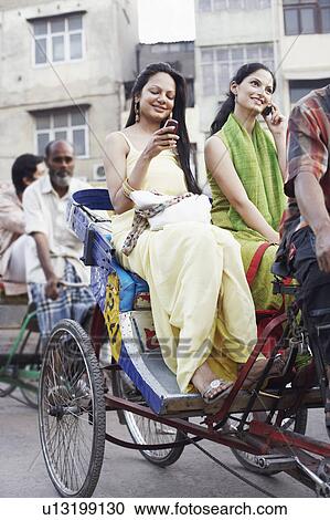 Stock Image - Two young women sitting in a rickshaw and using mobile phones. Fotosearch