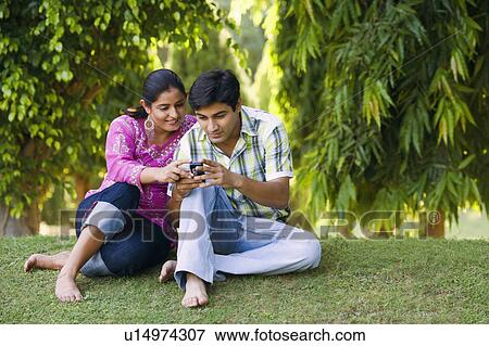 Young couple sitting in a lawn and looking at a mobile phone View Large Photo Image Stock Photo - Young couple sitting in a lawn and looking at a mobile phone. Fotosearch