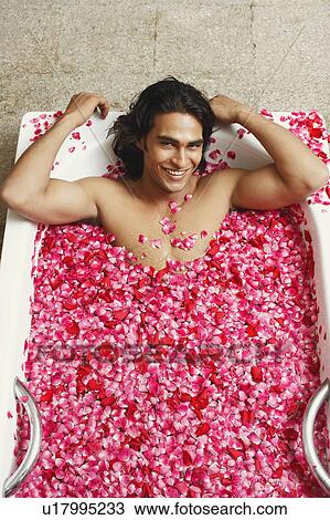 Stock Image - Portrait of a young man lying in a bathtub filled with rose petals and smiling. Fotosearch