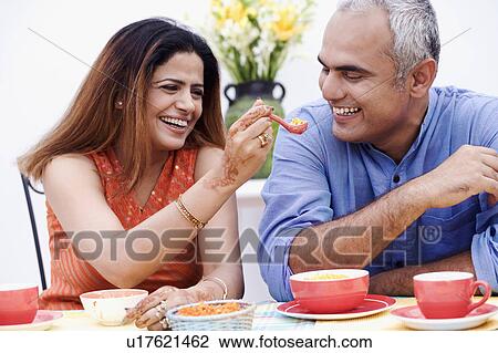 Stock Image - Mid adult woman feeding a spoonful of snacks to a mid adult man. Fotosearch
