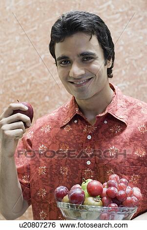 Stock Photograph - Portrait of a young man holding a bowl of fruits and smiling. Fotosearch