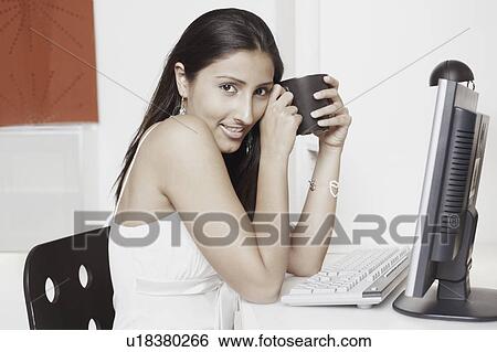 Portrait of a young woman sitting in front of a computer and holding a mug View Large Photo Image Stock Photograph - Portrait of a young woman sitting in front of a computer and holding a mug. Fotosearch