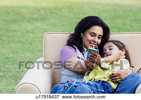 Stock Photo - Mid adult woman listening to an MP3 player with her daughter lying on her lap. Fotosearch