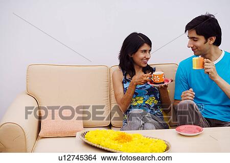 Picture - Young couple sitting on a couch and holding cups of tea. Fotosearch