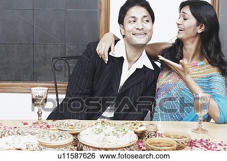 Stock Photograph - Young couple with arm around sitting at the dining table. Fotosearch