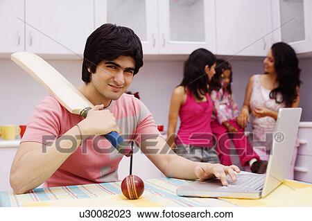 Young man holding a cricket bat and using a laptop in the kitchen View Large Photo Image Stock Image - Young man holding a cricket bat and using a laptop in the kitchen. Fotosearch