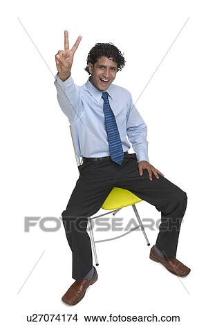 Picture - Portrait of a young man sitting on a chair and making a peace sign. Fotosearch