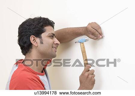 Close-up of a young man hammering a nail into the wall with a hammer View Large Photo Image Stock Photo - Close-up of a young man hammering a nail into the wall with a hammer. Fotosearch