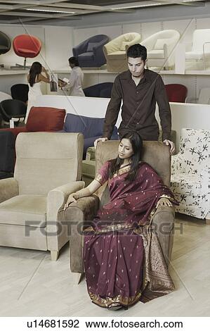 Stock Image - High angle view of a young woman sitting in an armchair in a furniture store with a young man standing behind her. Fotosearch