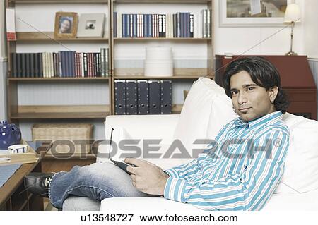 Stock Photo - Portrait of a young man sitting on a couch and holding a book. Fotosearch