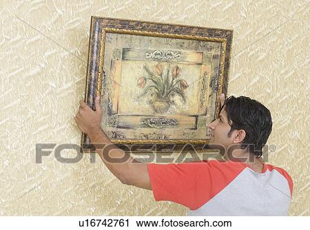 Stock Image - Rear view of a young man hanging a painting on a wall. Fotosearch