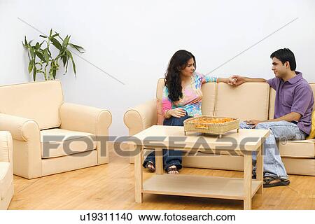Stock Image - Side profile of a young couple sitting on a couch and looking at each other. Fotosearch