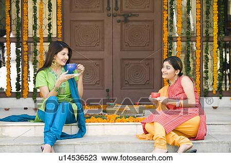 Stock Image - Two young women sitting at doorstep of a house and holding cups of tea. Fotosearch