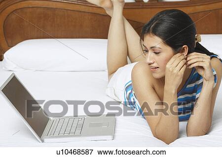 Stock Photo - Young woman lying on the bed and looking at a laptop. Fotosearch