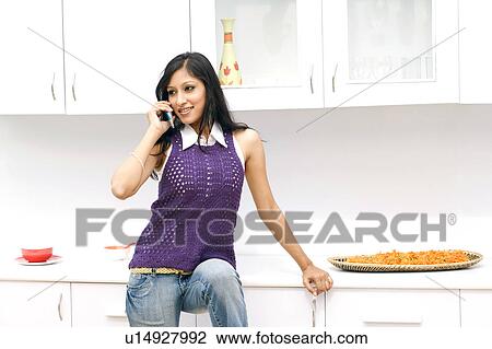 Stock Image - Young woman talking on a mobile phone in the kitchen. Fotosearch
