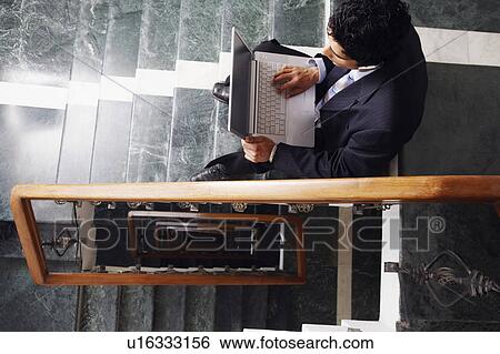 Stock Photograph - High angle view of a businessman sitting on the staircase and using a laptop. Fotosearch