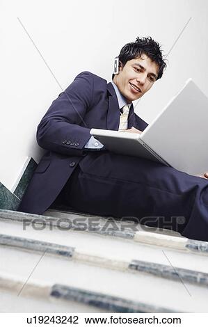 Stock Image - Low angle view of a businessman sitting on the staircase and using a laptop. Fotosearch