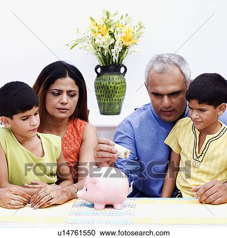 Stock Image - Mid adult man putting an Indian currency note into a piggybank with his wife and two sons beside him. Fotosearch
