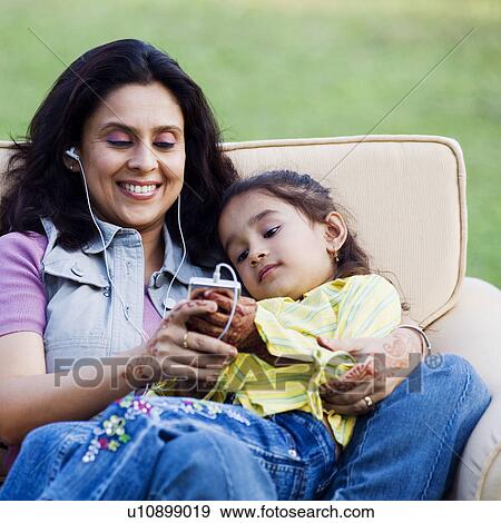 Stock Photo - Mid adult woman listening to an MP3 player with her daughter lying on her lap. Fotosearch