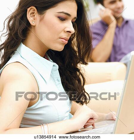 Side profile of a young woman using a laptop with a young man talking on a mobile phone in the background View Large Photo Image Picture - Side profile of a young woman using a laptop with a young man talking on a mobile phone in the background. Fotosearch