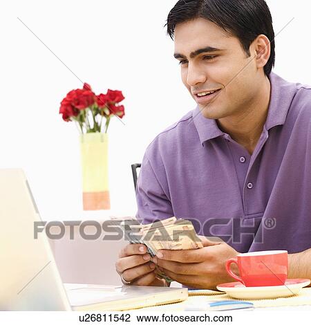 Young man sitting in front of a laptop and counting currency notes View Large Photo Image Stock Image - Young man sitting in front of a laptop and counting currency notes. Fotosearch