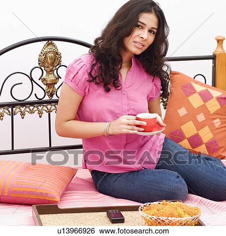 Stock Photograph - Young woman sitting on the bed and holding a cup of tea. Fotosearch
