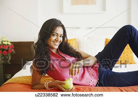 Stock Image - Portrait of a young woman lying on the bed and holding a pear. Fotosearch