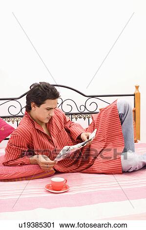 Stock Image - Young man reclining on the bed and reading a newspaper. Fotosearch