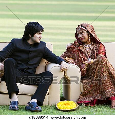 Picture - Newlywed couple in traditional wedding dress sitting in armchairs in a lawn and looking at each other. Fotosearch