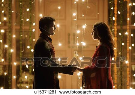 Stock Image - Side profile of a young couple standing face to face and holding each other's hands. Fotosearch