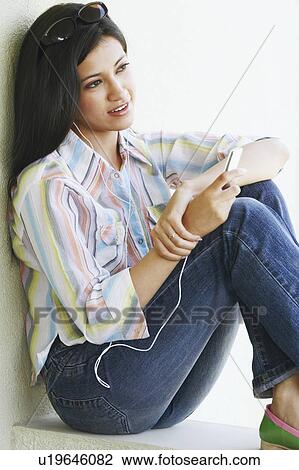 Stock Image - Side profile of a young woman sitting on a ledge and listening to an MP3 player. Fotosearch