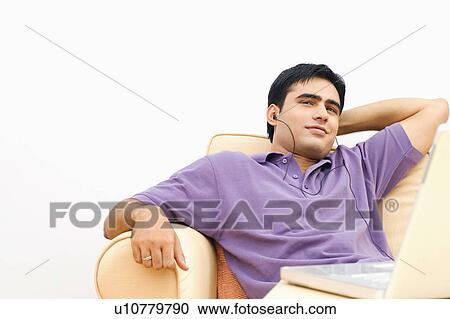Stock Image - Young man sitting on a couch in front of a laptop and listening to music. Fotosearch