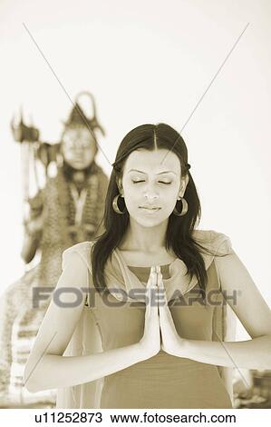 Stock Image - Close-up of a young woman standing in a prayer position in front of an idol of Lord Shiva. Fotosearch
